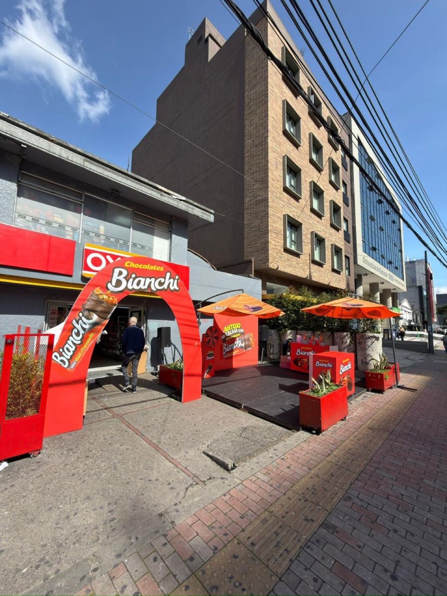 Arco de Chocolates Bianchi en la entrada de una tienda OXXO en Bogotá con terraza de sombrillas y materas de marca